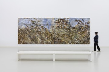 Ausstellungsraum mit einer weißen Bank und einer Besucherin vor einem querformatigen Gemälde von Anselm Kiefer, das ein Feld mit Ähren und Gräsern zeigt.
