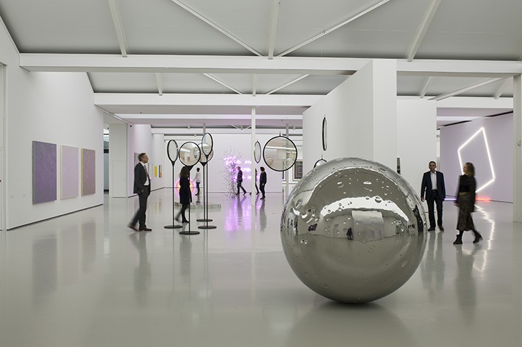 Die Ausstellungsansicht zeigt Besucherinnen und Besucher, die sich im Raum zwischen verschiedenen Skulpturen bewegen. Im Vordergrund ist eine große silberne Kugel zu sehen, die an einen Mond erinnert.