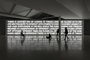 Bild 2: Im abgedunkelten Ausstellungsraum sind mehrere Besucher vor einem Lichtkunstwerk zu sehen, das sich im Boden spiegelt. Über mehrere Zeilen verläuft ein Satz aus Neonbuchstaben, der mit folgenden Worten beginnt: There is no other way of looking at things