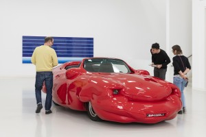 Bildergalerie: Bild 1: Drei Besucher betrachten aus unmittelbarer Nähe einen roten Porsche, der wie aufgeblasen wirkt.